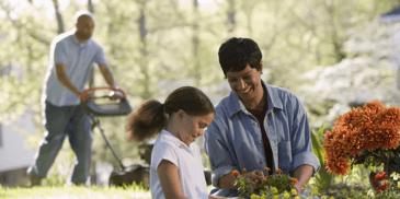 A woman and a child smiling and a man mowing the lawn in background